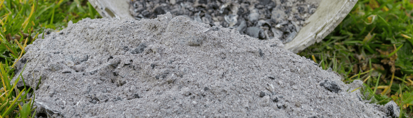 Haufen aus Holzasche auf grünem Rasen, im Hintergrund ein umgekippter Eimer mit Ascheresten.